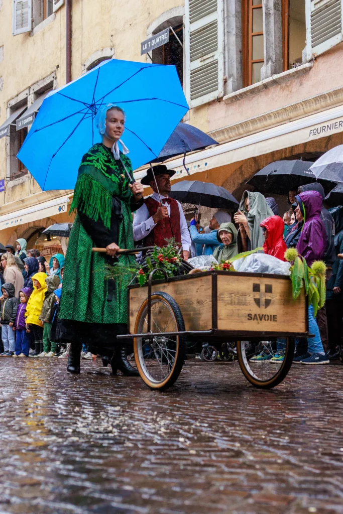 Femme en costume traditionnel savoyard sous son parapluie à Annecy avec une charrette de légumes
